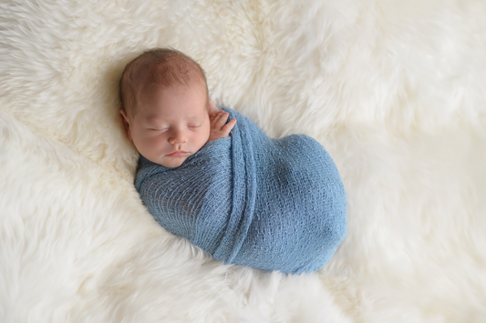 Baby wrapped in a blue swaddle blanket laying on a bed with white fur blanket